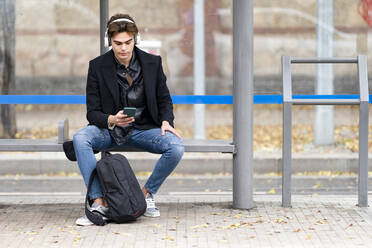 Handsome young man listening music while using smart phone sitting on bench at bus stop - GGGF00544
