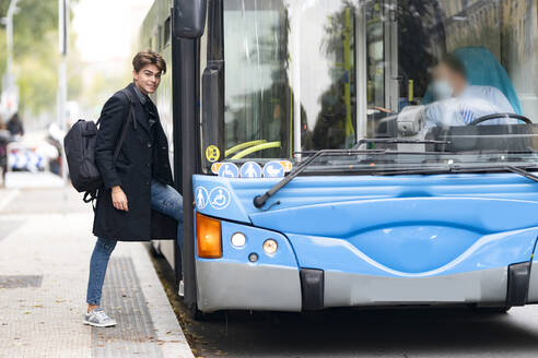 Lächelnder gut aussehender junger Mann mit Rucksack beim Einsteigen in einen Bus in der Stadt - GGGF00547