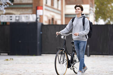 Smiling fashionable young man with backpack wheeling bicycle on footpath in city - GGGF00552