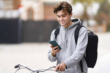 Smiling fashionable young man using smart phone while wheeling bicycle in city - GGGF00553