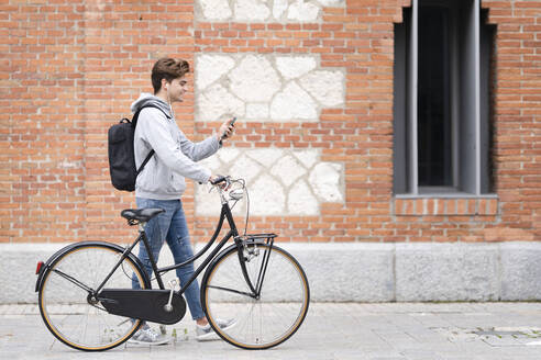 Smiling man on video call wheeling bicycle by building - GGGF00554