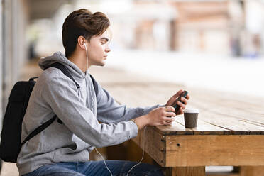 Gut aussehender Mann, der Musik hört, während er sein Smartphone in einem Straßencafé benutzt - GGGF00556