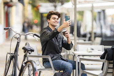 Lächelnder gut aussehender Mann, der ein Selfie mit einer Kaffeetasse durch ein Smartphone macht, während er in einem Straßencafé in der Stadt sitzt - GGGF00557