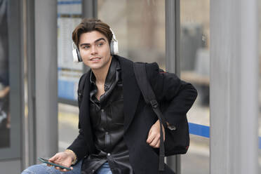 Hübscher junger Mann hört Musik und schaut weg, während er an der Bushaltestelle sitzt - GGGF00559