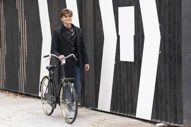 Lächelnder gut aussehender Mann, der auf dem Fußweg an der Mauer Fahrrad fährt - GGGF00562