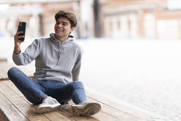 Fröhlicher junger Mann, der ein Selfie macht, während er auf einem Holztisch in einem Straßencafé in der Stadt sitzt - GGGF00568
