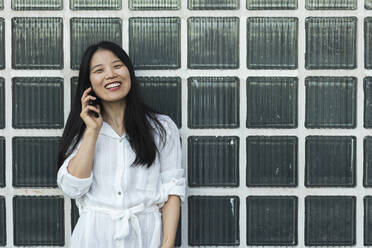 Smiling woman talking on mobile phone while standing against glass wall - PNAF00361