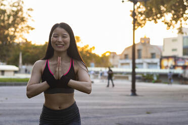 Smiling woman with hands clasped exercising in park during sunset - PNAF00368