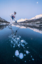 Eishockeyspieler beim Schlittschuhlaufen auf dem mit Eisblasen bedeckten Silsersee in der Abenddämmerung, Engadin, Kanton Graubünden, Schweiz, Europa - RHPLF18949