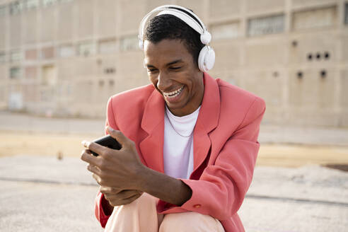 Glücklicher Mann mit Kopfhörern, der im Freien sitzend ein Mobiltelefon benutzt - AFVF07894