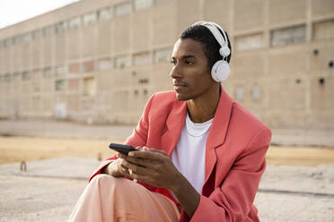 Junger Mann mit Kopfhörern und Mobiltelefon, der wegschaut, während er an einem Gebäude sitzt - AFVF07897