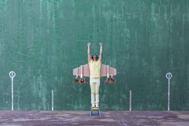 Fröhlicher Mann mit handgefertigten Flügeln aus Pappe, der auf einem kleinen Stuhl mit erhobenen Armen steht und nach oben schaut, während er vom Erfolg träumt - ADSF19500