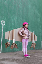 Von unten Seitenansicht von niedlichen Mädchen in Pilot Helm und handgemachte kreative Karton Flügel stehen auf der Straße auf grünem Hintergrund wegschauen - ADSF19510
