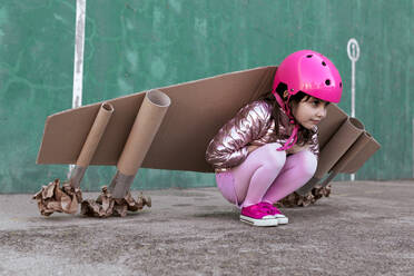 Seitenansicht eines niedlichen kleinen Mädchens mit Flugzeugflügeln aus Pappe und Schutzhelm, das auf der Straße sitzt und wegschaut - ADSF19511