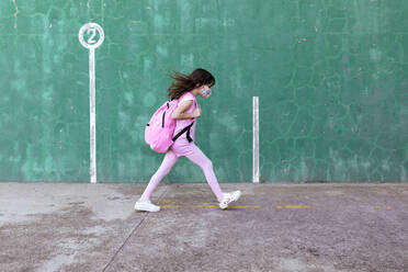 Side view of schoolgirl with rucksack and in protective mask walking along street during coronavirus epidemic - ADSF19519