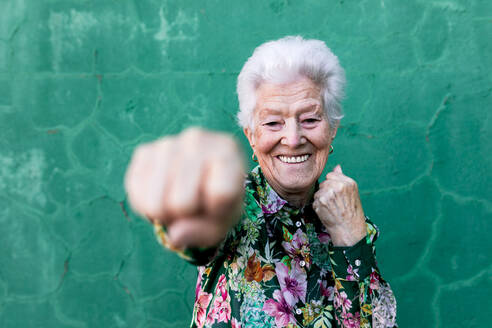Fröhliche ältere grauhaarige Dame in stilvoller bunter Bluse, die Spaß hat und in die Kamera boxt, während sie an einer grünen Wand steht - ADSF19522