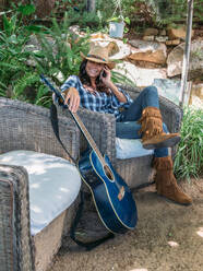 Positive erwachsene ethnische Frau in stilvollem Cowboy-Outfit, die auf einem Smartphone spricht, während sie in einem Korbsessel neben einer Gitarre auf einer Gartenterrasse mit grünen Topfpflanzen sitzt - ADSF19544