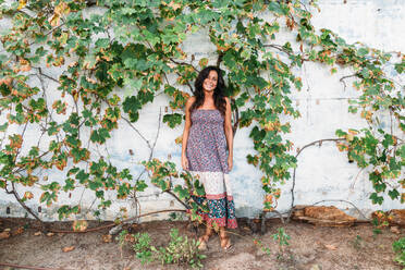 Full length of smiling adult ethnic female in casual summer dress standing against weathered stone wall with green ivy plants - ADSF19546