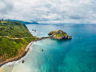 Drohnenansicht eines gepflasterten Weges, der über eine Steinbrücke und den Kamm eines felsigen Hügels zu einem einsamen Haus auf der Insel Gaztelugatxe führt, umgeben von ruhigem Meerwasser unter bewölktem Himmel im Baskenland - ADSF19559