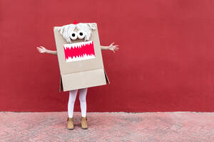 Unrecognizable playful kid wearing funny costume of monster made of carton box standing on street during holiday celebration - ADSF19575