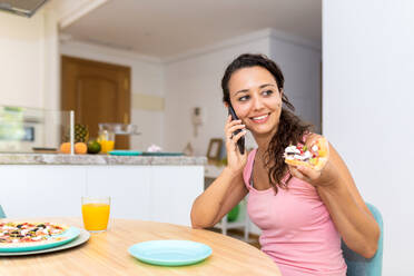 Happy young female in casual wear answering phone call while sitting at table and eating pizza during lunch at home - ADSF19576