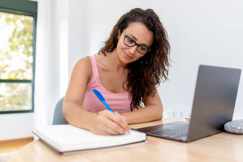 Positive junge Studentin in Freizeitkleidung und mit Brille sitzt am Tisch mit Laptop und schreibt Informationen in ein Heft während des Online-Studiums zu Hause - ADSF19577