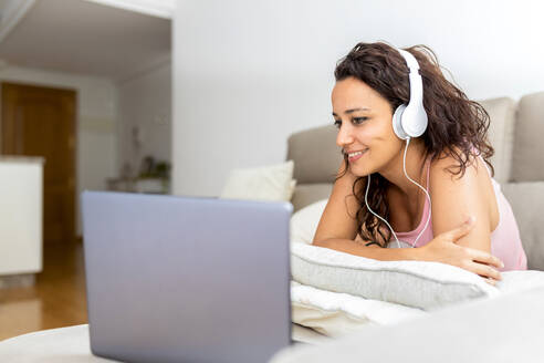 Cheerful relaxed female lying on sofa with laptop and enjoying movie in headphones while chilling at weekend at home - ADSF19578