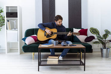 Junger Musiker spielt Gitarre, während er auf dem Sofa im Studio sitzt - GIOF10175