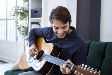 Lächelnder Gitarrist spielt Gitarre, während er auf dem Sofa im Studio sitzt - GIOF10176
