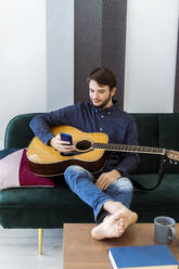 Gitarrist, der ein Mobiltelefon benutzt, während er mit seiner Gitarre auf dem Sofa im Studio sitzt - GIOF10181