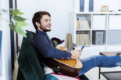 Lächelnder Musikprofi, der ein Mobiltelefon benutzt, während er auf dem Sofa im Studio sitzt - GIOF10182