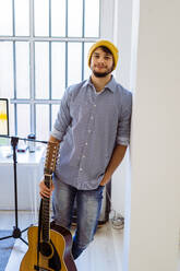 Lächelnder Gitarrist mit gestricktem Haar, stehend mit Gitarre im Studio - GIOF10200