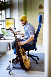 Lächelnder Musiker, der die Gitarre nimmt, während er am Schreibtisch im Studio sitzt - GIOF10211