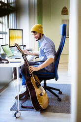 Smiling musician taking guitar while sitting at desk in studio - GIOF10211