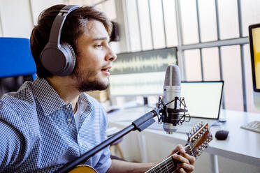 Männlicher Sänger mit Kopfhörern und Mikrofon singt, während er im Aufnahmestudio Gitarre spielt - GIOF10233