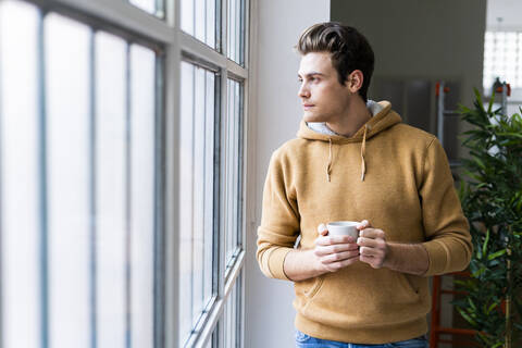 Young man with coffee cup contemplating while looking through window in new apartment stock photo