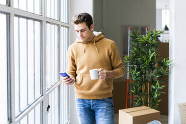 Junger Mann mit Kaffeetasse und Smartphone am Fenster eines neuen Hauses - GIOF10273