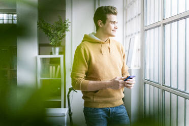 Lächelnder Mann schaut durch ein Fenster und hält ein Handy in seinem neuen Zuhause - GIOF10276