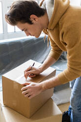 Young man writing on cardboard box while moving into new loft apartment - GIOF10287
