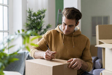 Young man writing on cardboard box with pen in new home - GIOF10288