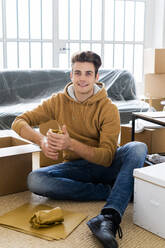 Lächelnder Mann, der ein eingewickeltes Trinkglas in einer Schachtel in seiner neuen Loftwohnung verpackt - GIOF10293