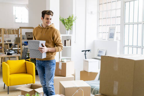 Smiling young man holding boxes while standing in new loft apartment - GIOF10294