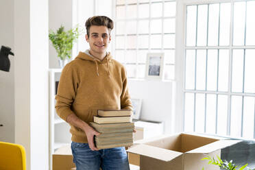 Lächelnder junger Mann mit Büchern in einem Karton in seiner neuen Loftwohnung - GIOF10298