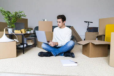 Junger Mann, der einen Laptop benutzt, während er auf dem Boden in einem Raum mit Kisten in einer neuen Wohnung sitzt - GIOF10299