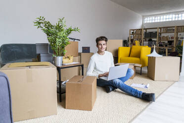 Lächelnder junger Mann mit Laptop in einem unordentlichen Wohnzimmer in einer neuen Wohnung - GIOF10302