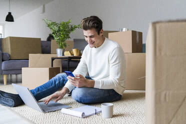 Lächelnder Mann mit Laptop, der ein Smartphone benutzt, während er im Wohnzimmer seines neuen Hauses sitzt - GIOF10305