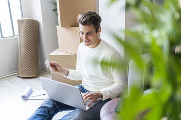 Lächelnder junger Mann mit Kreditkarte und Laptop beim Umzug in eine neue Wohnung - GIOF10308