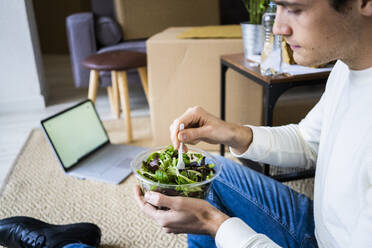 Young man with laptop having salad in living room of new loft apartment - GIOF10310