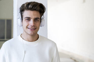 Lächelnder junger Mann hört Musik über Kopfhörer an einer Säule in seinem neuen Haus - GIOF10321