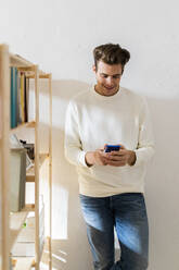 Smiling young man using smart phone against white wall by bookshelf at home - GIOF10325